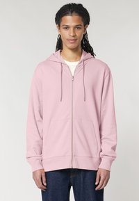 Svetlo roza ženska zip-up hoodie s kapuco na vrvico, sprednjimi žepi in sproščenim krojem, v kombinaciji s temno modrimi kavbojkami. Gladka tekstura.