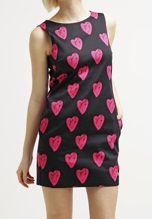 Černé šaty bez rukávů s jasně růžovými srdcovými vzory, nošené osobou s jednou rukou v kapse a druhou volně podél těla.