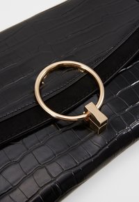 Pochette en cuir noir embossé effet croco avec un grand anneau circulaire doré et une fermeture rectangulaire sur le rabat avant.