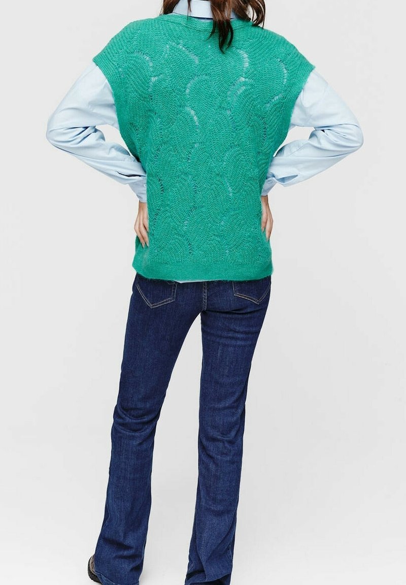 Personne portant un pull sans manches tricoté bleu sarcelle sur une chemise bleu clair et un jean bleu foncé, debout les mains sur les hanches, dos tourné vers le spectateur.
