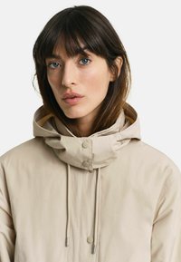Beige hættetrøje med snoretræk i halsen, trykknaplukninger og glat overflade. Har et diskret design uden yderligere mønstre eller detaljer.