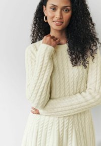 Pull en maille torsadée couleur crème, avec un col rond et des manches longues, doté d'un motif texturé sur l'ensemble du tissu.