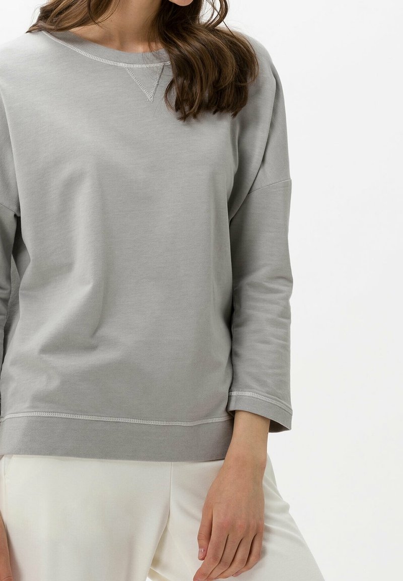 Femme portant un sweat-shirt gris clair à manches longues avec des poignets et un ourlet côtelés, associé à un pantalon blanc sur un fond blanc uni.
