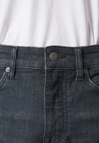 Zone de la taille d'un jean en denim bleu foncé avec bouton et passants de ceinture porté avec une chemise blanche rentrée.