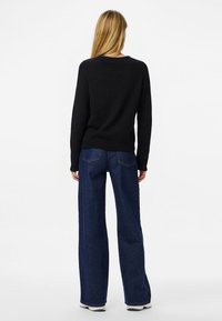 Hosszú szőke hajú nő háttal áll, fekete pulóvert, bő szárú sötétkék farmert és fehér tornacipőt visel fehér háttér előtt.