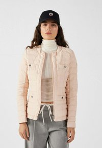 Giacca trapuntata rosa chiaro con colletto alto, due tasche frontali e dettagli metallici argentati. Indossata sopra un dolcevita bianco e pantaloni grigi.