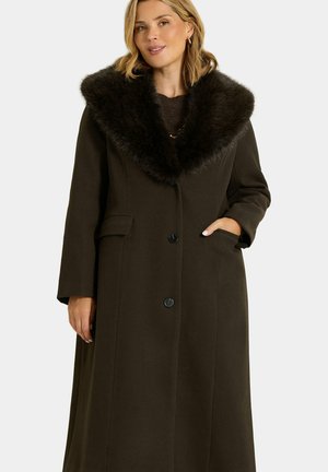 Femme portant un long manteau marron foncé avec un grand col en fourrure noire, deux boutons, et sa main droite dans la poche sur un fond uni.