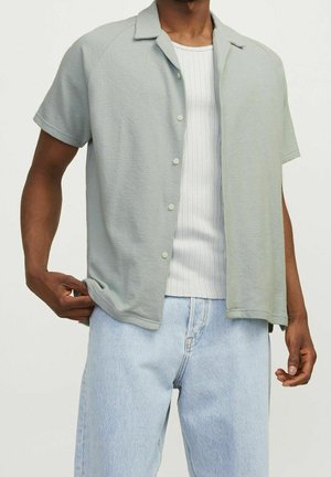 Mand iført en lysegrå kortærmet skjorte med knapper over en hvid ribbet tanktop og lyseblå denimjeans.