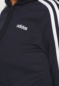 Marinblå zip-up hoodie i bomullsmix, med vita accentränder längs ärmarna och en broderad logotyp på bröstet.