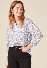 Blusa bianca di cotone con motivo astratto blu, con colletto e maniche a tre quarti; abbinata a pantaloni di denim neri.