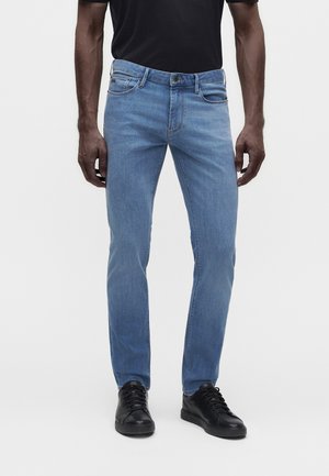 Mand iført lyseblå slim-fit jeans, sort skjorte og sorte sneakers, stående mod en ensfarvet lys baggrund.