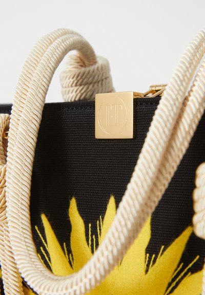 Canvas bag with a black body, yellow floral design, and cream rope handles. Features a gold logo plaque at the top.