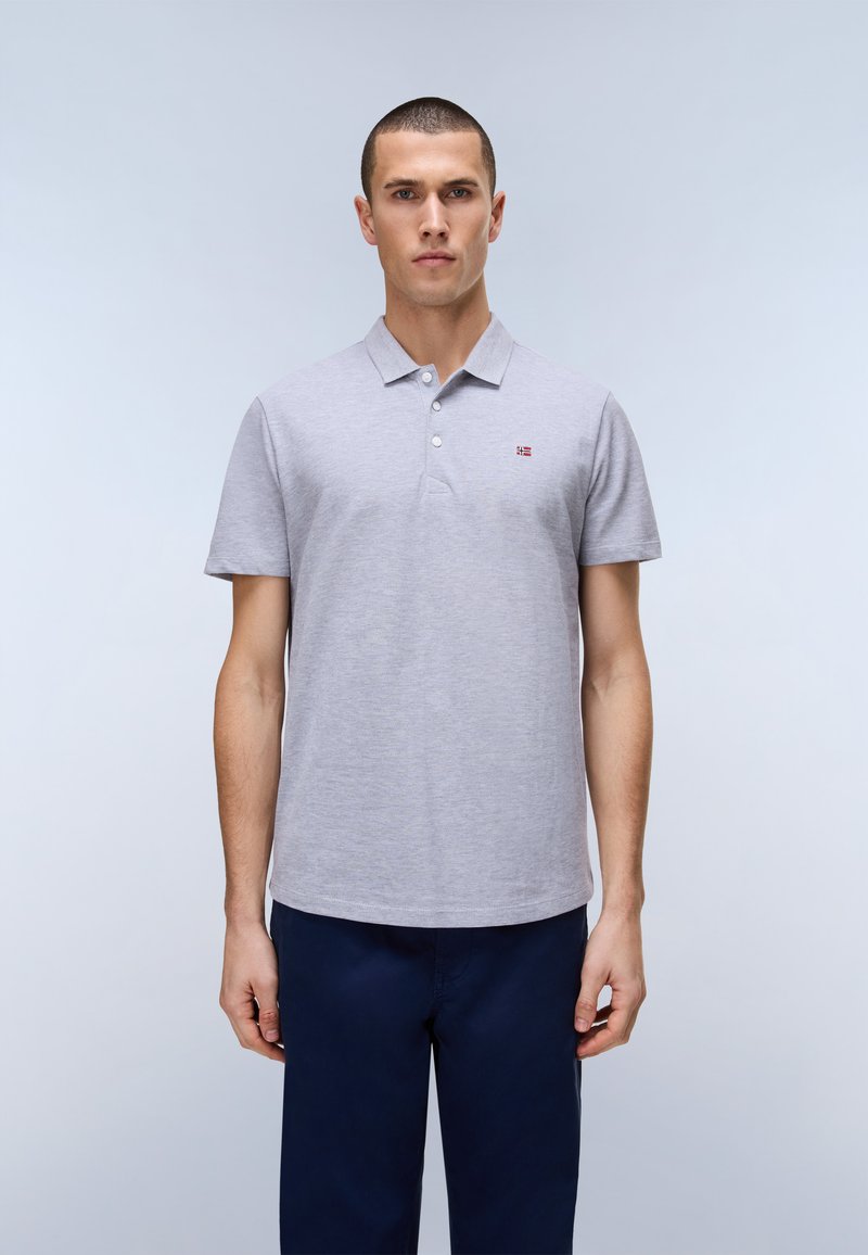 Uomo che indossa una polo a maniche corte grigio chiaro con un piccolo logo e pantaloni blu scuro, in piedi contro uno sfondo chiaro e uniforme.