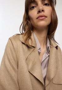 Giacca in suede beige con colletto a punta, morbida al tatto e dal fit ampio, indossata sopra una blusa in satin grigio chiaro. Dettagli di cucitura sottili.