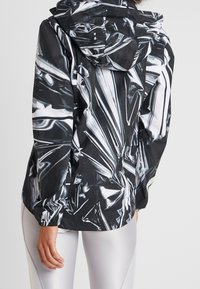 Windbreaker jacket in een zwart-wit reflecterend materiaal. Voorzien van een capuchon, elastische mouwen en een abstracte patroon dat lijkt op gekreukte folie.