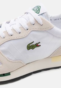Sneaker med en blanding av hvit nylon og beige semsket skinn, med en grønn krokodillelogo, hvite lisser og en kontrasterende svart gummisåle.