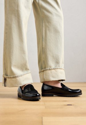 Person, der bequeme beigefarbene Hose mit Aufschlägen und glänzende schwarze Leder-Slipper trägt, steht auf einem Holzboden vor einem hellen Hintergrund.