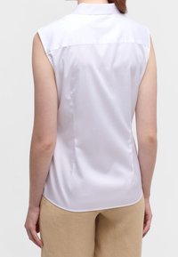 Blouse blanche sans manches avec col pointu, texture satinée lisse et coutures latérales, portée avec un pantalon beige clair.