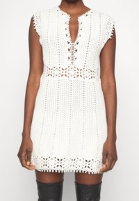 Abito in crochet bianco con scollatura a V profonda, maniche corte a cappuccio e dettagli in pizzo intricati lungo l'orlo e la vita. Motivo testurizzato in tutto l'abito.
