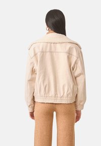 Chaqueta de algodón beige con un cuello esponjoso, dobladillo elástico y puños. La parte trasera es sencilla con detalles de costura. Se lleva sobre pantalones marrones ajustados.