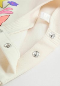 Cremefarbener Stoff mit silbernen Druckknöpfen und teilweisem mehrfarbigem Blumendesign auf einem weichen, gestrickten Kleidungsstück.