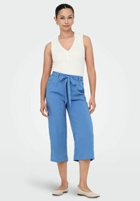 Haut blanc sans manches à côtes avec col en V et détail de boutons ; pantalon court bleu avec taille nouée et poches avant ; ballerines beige.