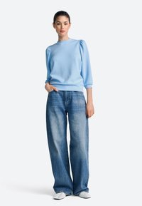 hele sinine, kubjakatel varrukatel pullover-kampsun, kombineeritud kõrge vöökohaga, laiade säärega siniste teksadega. Modell seisab, üks käsi puusal.