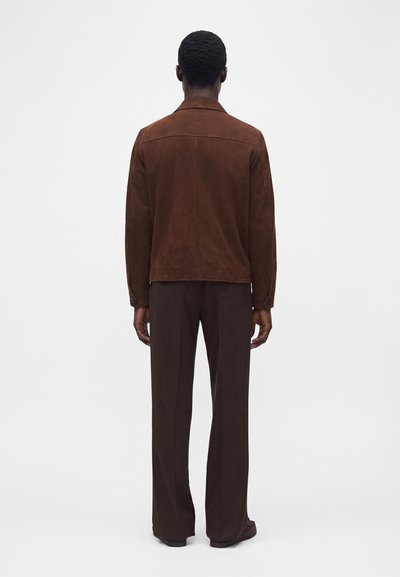 Homme debout, dos tourné, portant une veste en daim marron et un pantalon marron foncé sur un fond blanc uni.