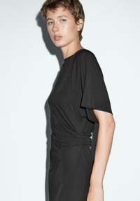 Vestito nero con una vestibilità rilassata, maniche corte e dettaglio arricciato in vita. Texture liscia e design minimalista.