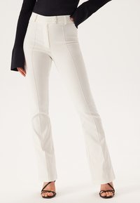 Pantalons blancs évasés à taille haute, dotés de doubles boutons et de coutures avant. Tissu lisse avec un léger brillant, associés à des talons noirs à lanières.