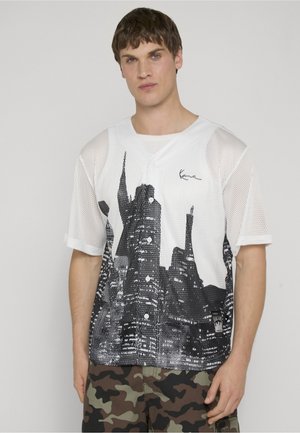 Joven con una camisa blanca de malla abotonada con un estampado negro de paisaje urbano y pantalones de camuflaje, de pie frente a un fondo sencillo.