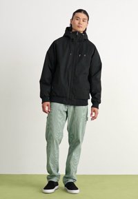 Black zip-up jacket with a hood, elastic cuffs, and front pockets. Paired with green striped pants and black slip-on shoes.