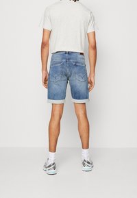 Mies joka on pukeutunut vaaleanharmaaseen t-paitaan, ylös käärittyihin sinisiin farkkushortseihin, valkoisiin sukkiin ja hopeisiin juoksukenkään, seisoo valkoista taustaa vasten.