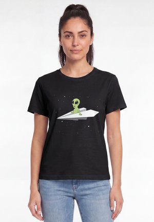 Femme portant un t-shirt noir avec un extraterrestre vert en dessin animé assis sur un avion en papier blanc dans un design spatial, associée à un jean bleu clair.