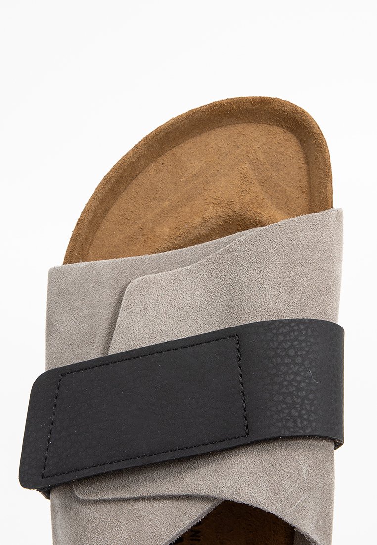 Grijze suède sandaal met een zwarte gestructuurde band. Gecontourde bruine binnenzool met een gladde afwerking, ontworpen voor comfort en ondersteuning.