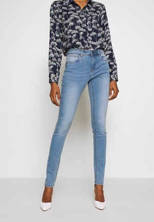 Woman wearing high-waisted light blue skinny jeans, floral long-sleeve blouse, and white pointed-toe heels, standing against a plain background.