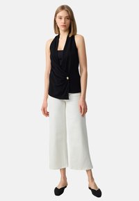 Top senza maniche nero con design a torsione e dettaglio in bottone dorato, abbinato a pantaloni bianchi a gamba larga e scarpe nere slip-on.