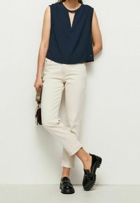 Top senza maniche blu navy con scollatura a chiave, abbinato a pantaloni slim-fit beige chiaro e mocassini neri spessi, reggendo una borsa in mano.