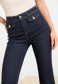 Mörkblå jeans i denintyg med hög midja, med guld detaljer och en klassisk femficksdesign. Texturen är slät med synliga sömmar.