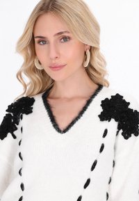 Weißer Pullover mit schwarzen floralen Applikationen an den Schultern, V-Ausschnitt und strukturierten Akzenten. Weiches Material mit lockerer Passform und dekorativen Nähten.