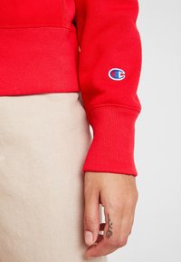 Rode sweatshirt met geribde mouwen, met een cirkelvormig blauw en wit logo op de linker mouw. Beige stof zichtbaar eronder.