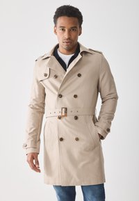 Abrigo trench beige con botones de doble abertura y cintura ceñida con cinturón. Presenta un cuello con muescas y dos bolsillos frontales. Textura de tela suave.