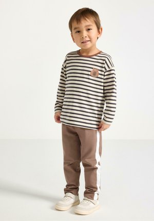 Nuori poika raidallisessa pitkähihaisessa paidassa, jossa on karhutasku, ruskeissa housuissa, joissa on valkoiset sivuraidat, ja valkoisissa tennareissa seisoo yksinkertaista taustaa vasten.