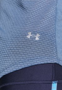 Detailní záběr na modré strukturované sportovní tričko s logem Under Armour nad tmavě modrým upnutým páskem sportovního oblečení.