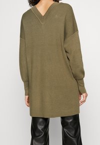 Pull en tricot vert olive avec un décolleté en V, des épaules tombantes et une coupe décontractée, présentant une texture côtelée et des manches longues. Associé à un pantalon noir.