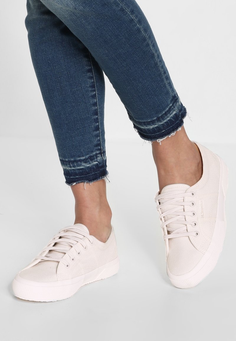 Personne portant un jean bleu foncé à ourlet effiloché et des baskets en toile écru à lacets, debout sur un sol gris clair uni.
