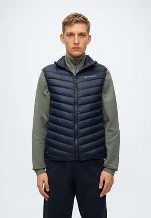 Jeune homme portant un gilet matelassé noir par-dessus une chemise grise à manches longues avec fermeture éclair, debout devant un fond blanc uni.