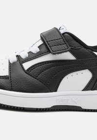 Schwarzer und weißer Sneaker mit einem Obermaterial aus synthetischem Leder, ausgestattet mit einem Klettverschluss, weißen Schnürsenkeln und einer strukturierten Gummisohle.