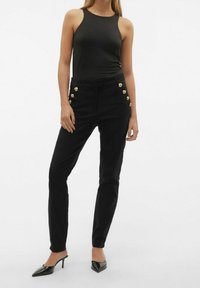 Pantalones negros ajustados con bolsillos laterales y acentos de botones dorados, combinados con una blusa negra sin mangas ajustada. Tela suave y piernas cónicas.