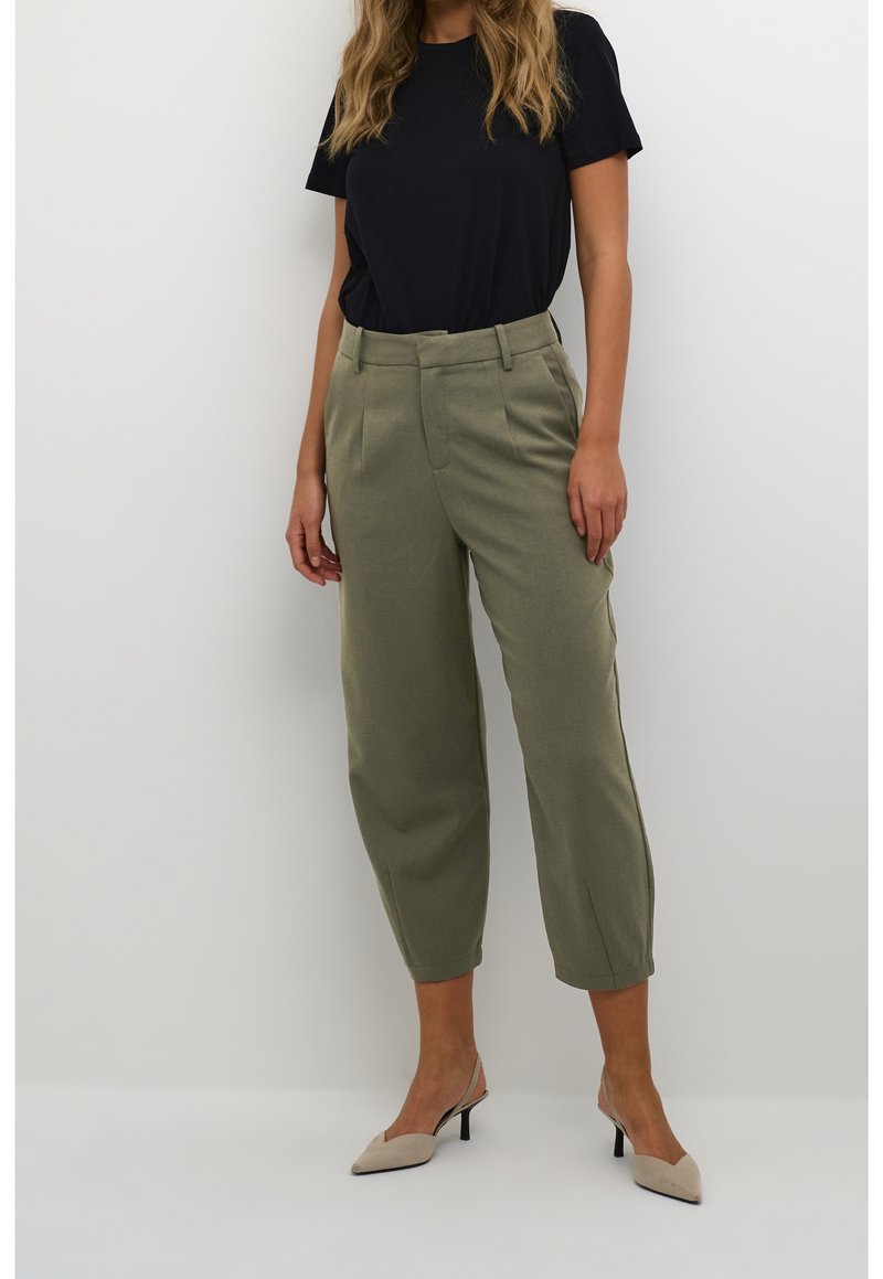 Femme portant un pantalon fuselé taille haute de couleur vert olive, un haut noir à manches courtes et des talons slingback beiges à bout pointu, debout contre un mur blanc.